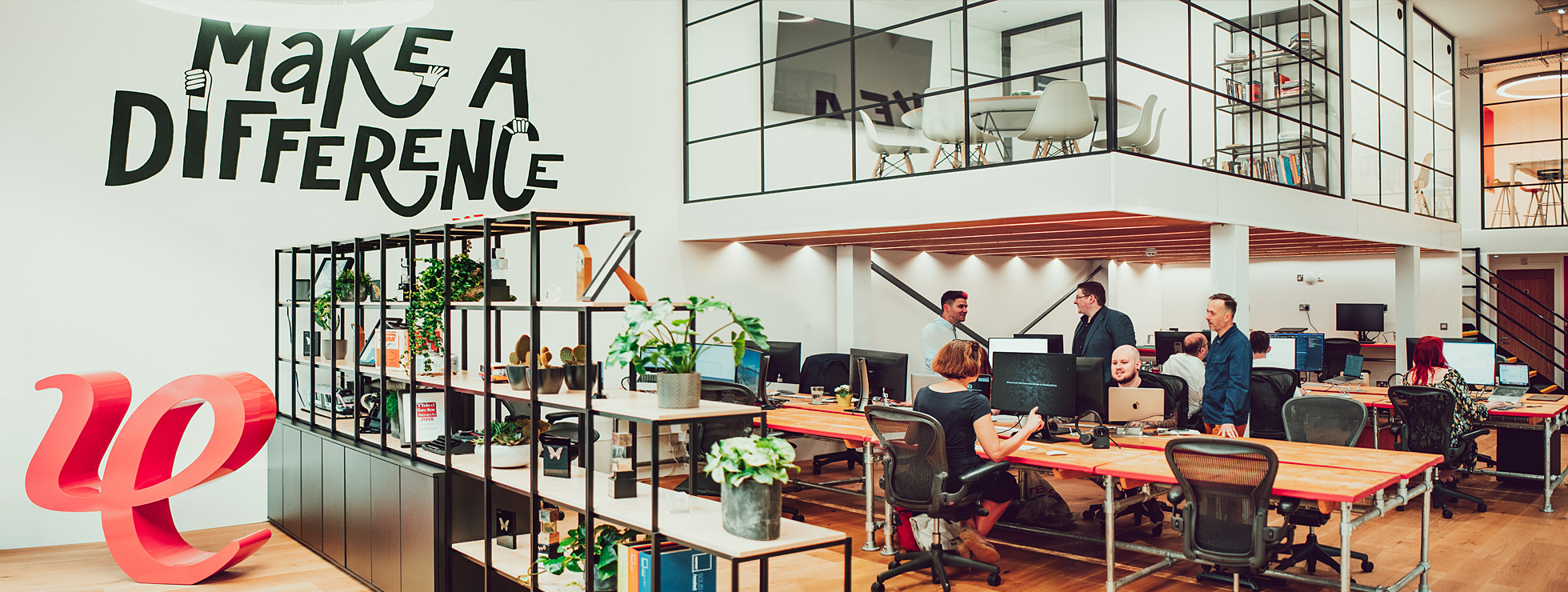 A view across the IE Brand studio in Birmingham, showing members of the agency team collaborating on projects. There's a mural on the wall with the agency's motto "Make a difference, est. 1994" and a large red 3D version of IE's logo marque.